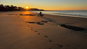 Une plage où l’on voit des empreintes de pas menant à l’eau, une personne à vélo et le soleil se couchant à l’horizon.
