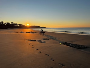 Ein Strand mit Fußspuren, die zum Wasser führen, eine Person fährt Fahrrad und die Sonne geht im Hintergrund unter.