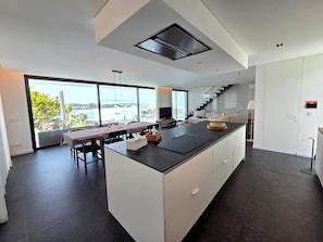 Eine moderne Küche mit einer großen Kochinsel, einem Essbereich mit Tisch und Stühlen sowie Blick durch große Fenster.