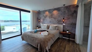 Un dormitorio moderno con una cama grande, vista al mar y una pared de concreto decorativa.