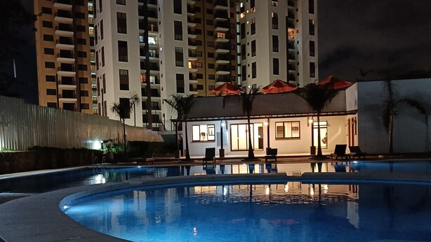 Ein Pool mit gewölbter Kante, umgeben von einer gefliesten Terrasse. Im Hintergrund sind mehrstöckige Wohngebäude mit Balkonen zu sehen.