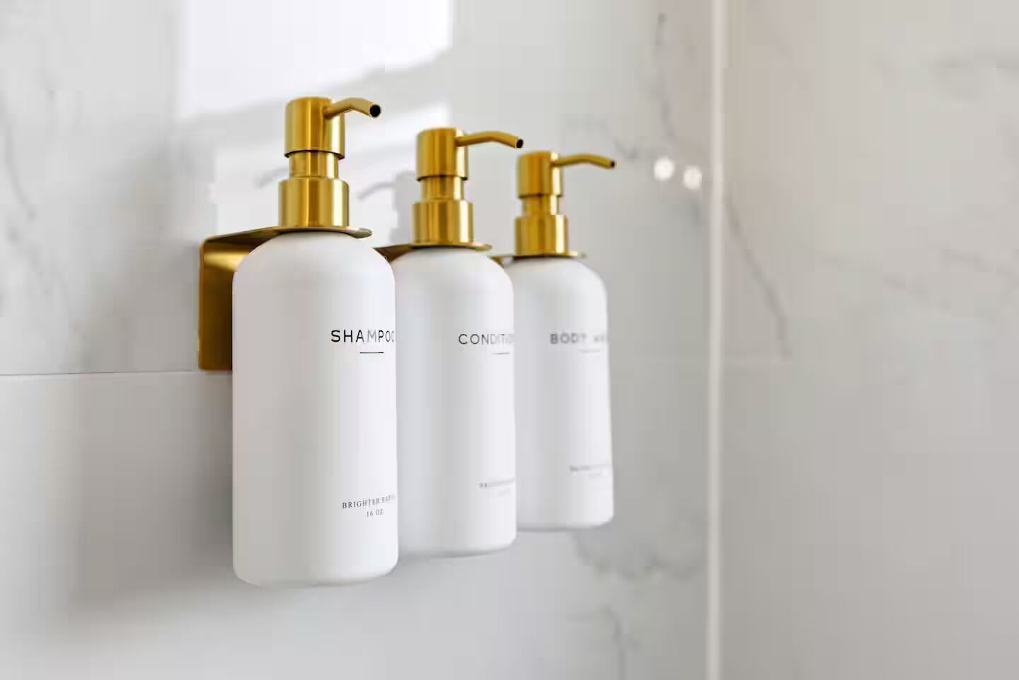 Three white bottles with gold pumps labeled 'SHAMPOO,' 'CONDITIONER,' and 'BODY WASH' mounted on a white tiled wall.