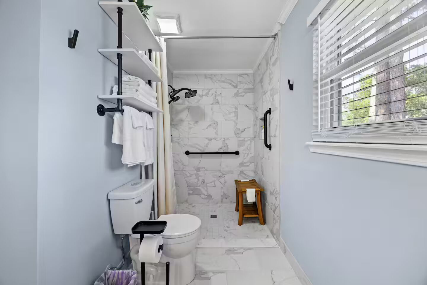 Un baño con inodoro, ducha, paredes de mármol, un taburete de madera pequeño y una ventana con persianas.
