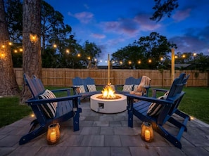 An outdoor seating area with a circular fire pit, surrounded by lounge chairs and string lights.