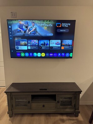 Televisão de tela plana fixada na parede, exibindo a interface Samsung TV Plus com diversos aplicativos de streaming.