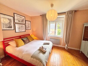 A bedroom with a wooden floor, a bed with white bedding, a red headboard, and a window with curtains. - Petite maison à l'orée du bois (Sillery)