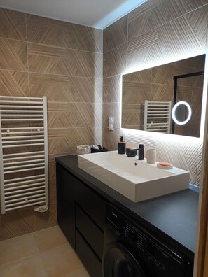 A modern bathroom with a large mirror, a white sink, a towel rack, and a washing machine. - Vrbo Property (Marseille)