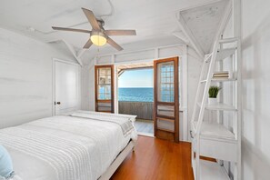 Un dormitorio con cama, ventilador de techo, piso de madera y vista al mar a través de puertas abiertas.