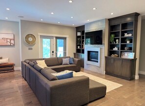A modern living room with a sectional sofa, a fireplace, built-in shelving, and a large window.