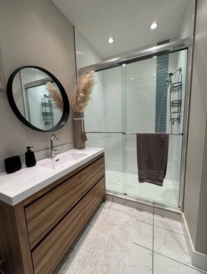 A modern bathroom with a glass shower enclosure, a round mirror, a wooden vanity, and a towel hanging on the shower door.