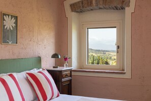 A bedroom with a bed, a nightstand, a lamp, a vase with flowers, and a window with a view of the countryside. - Il Poggione 10, Emma Villas (Crocino)