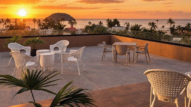Eine Dachterrasse mit Sitzgelegenheiten im Freien, Blick auf den Sonnenuntergang und Palmen.
