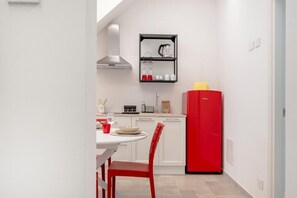 Une cuisine de taille réduite avec des meubles blancs, un réfrigérateur rouge et une étagère noire où sont rangés les ustensiles de cuisine.