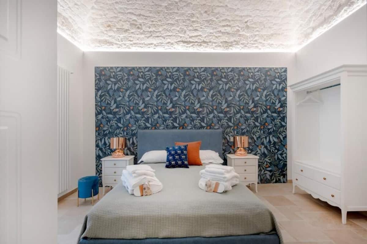 Un dormitorio con una cama, dos mesitas de noche, un aparador y una pared azul con estampado floral.