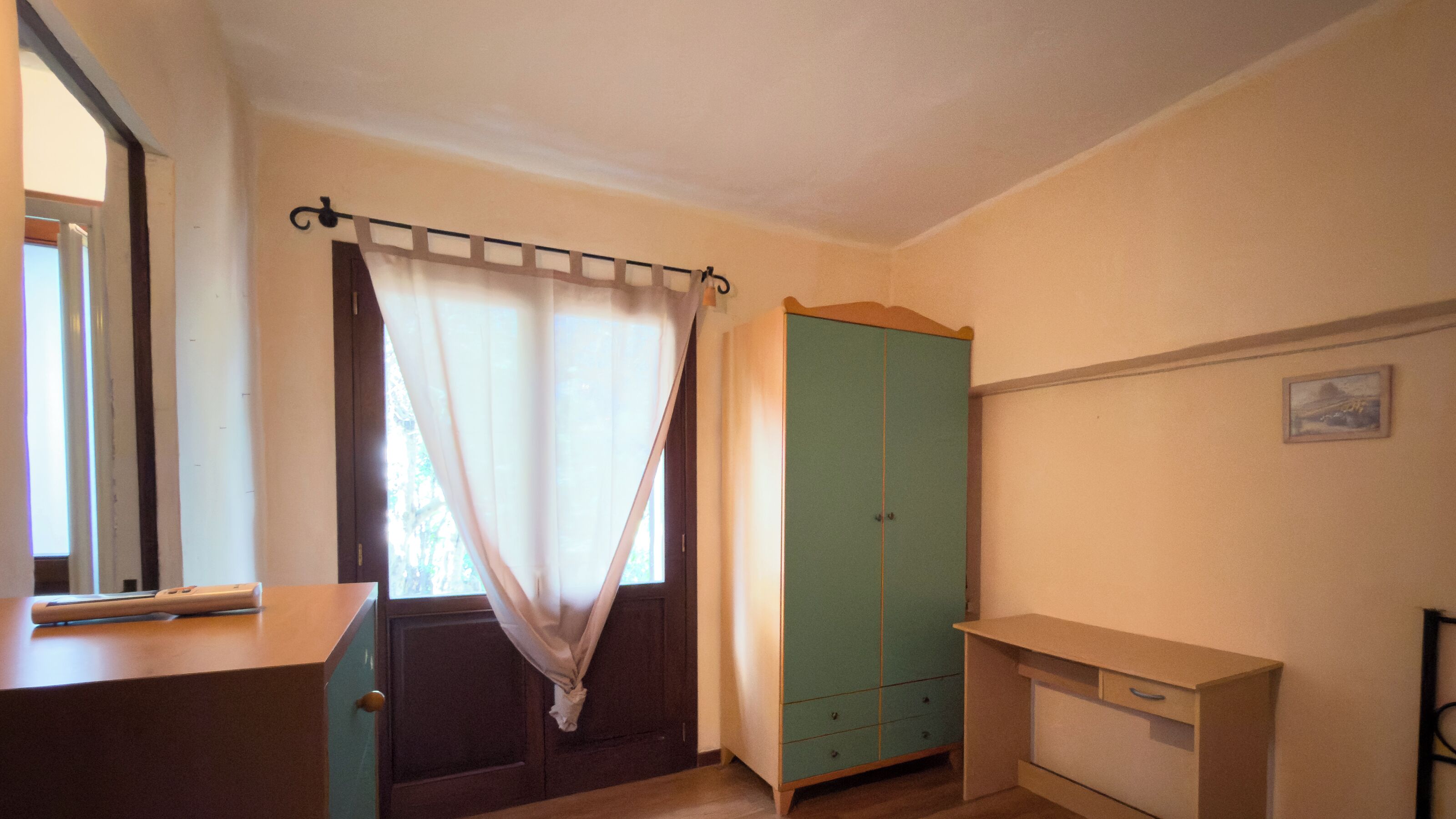 A room with a wooden desk, a green wardrobe, and a window with curtains.