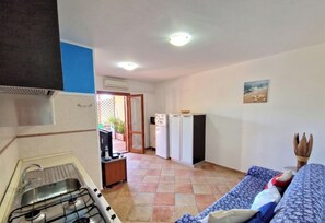 Une cuisine compacte équipée d’une cuisinière à gaz, d’un évier et d’un réfrigérateur. On y trouve un petit coin repas avec une table et des chaises, ainsi qu’un espace salon avec un canapé et une plante décorative.