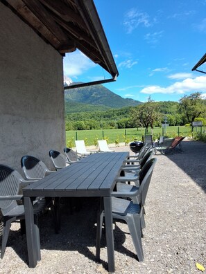 Eine Terrasse im Freien mit Tisch und Stühlen, einem Grill und Blick auf Berge und grüne Felder.