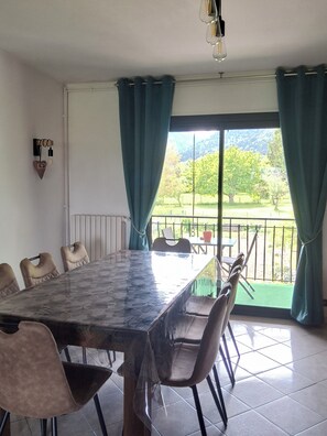 Ein Essbereich mit großem Tisch, Stühlen, Blick auf einen Balkon mit viel Grün und einer Wandleuchte.