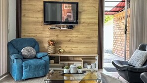 Ett vardagsrum med en tegelvägg, en väggmonterad TV, en blå soffa, en grå soffa och ett soffbord i glas.