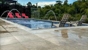 En pool med vattenstrålar, röda och beige solstolar samt en kakelbeklädd terrass.