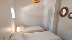Ein kleines, helles Schlafzimmer mit zwei Betten, einer Nachttischlampe, einem an der Wand befestigten Spiegel und einem Fenster, das natürliches Licht hereinlässt.