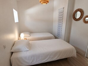 Ein kleines, helles Schlafzimmer mit zwei Betten, einer Nachttischlampe, einem an der Wand befestigten Spiegel und einem Fenster, das natürliches Licht hereinlässt.