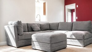 Ein modernes Wohnzimmer mit grauem Ecksofa und Hocker, einem großen Fenster und einer roten Akzentwand.