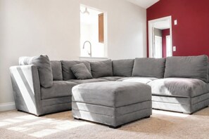 Ein modernes Wohnzimmer mit grauem Ecksofa und Hocker, einem großen Fenster und einer roten Akzentwand.