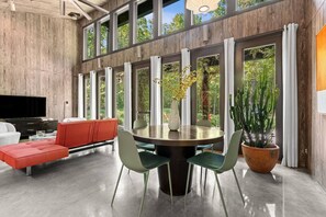 A modern living room with a round dining table, red sofa, and large windows offering a view of greenery. - Midnight Hollow in Fennville (Fennville)