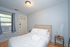 A bedroom with a bed, a bedside table, a lamp, a window with curtains, and a door.
