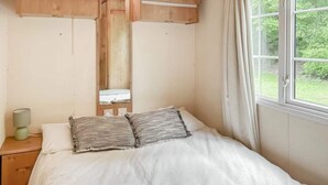 Ein Einzelbett mit weißer Matratze und zwei Kissen, ein Nachttisch aus Holz mit einer Lampe und ein Fenster mit Blick auf Grünflächen.