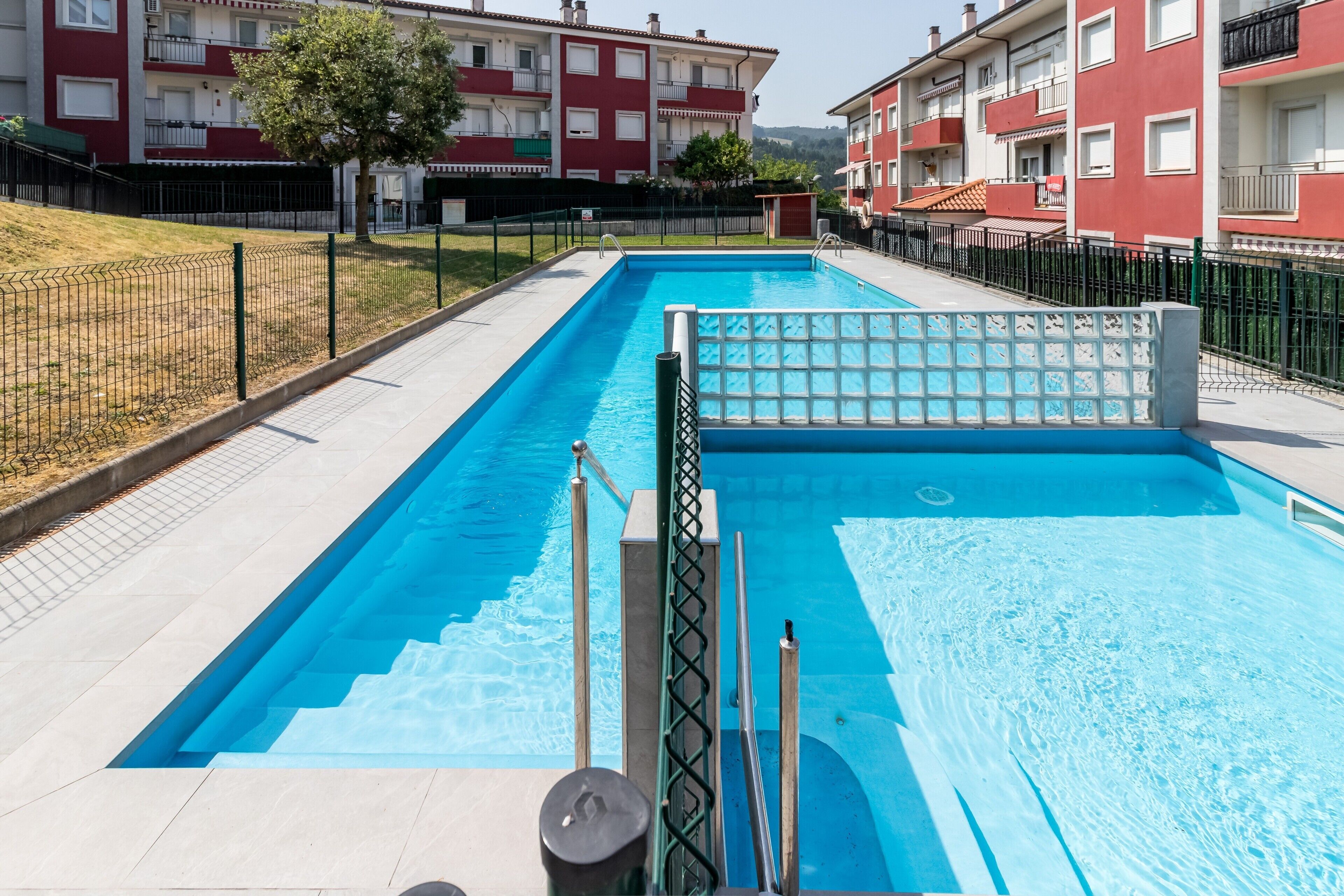 Una piscina rectangular amb terrassa enrajolada, envoltada per una tanca i edificis d'altes plantes a prop.