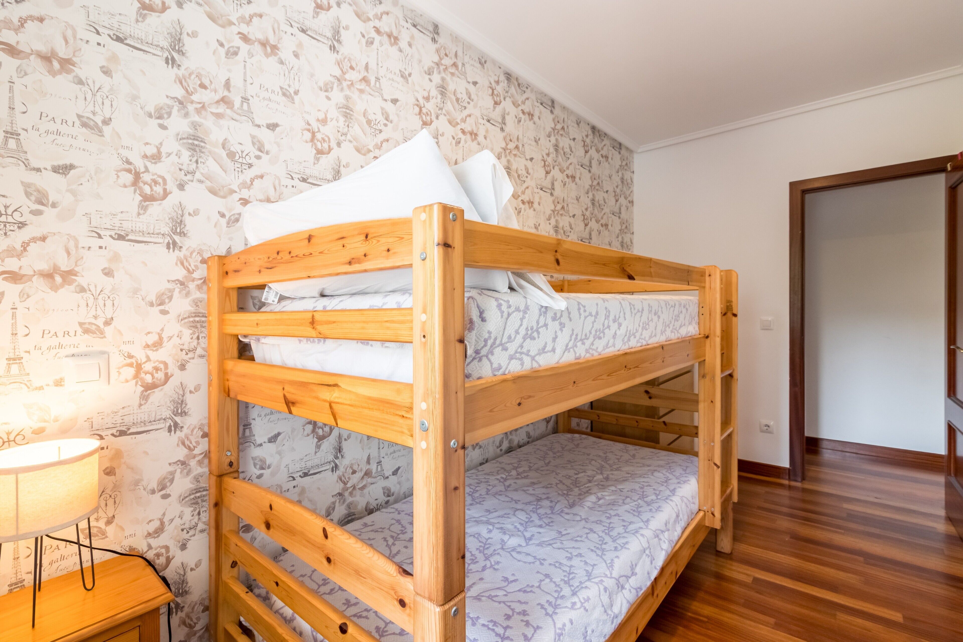 Una cama litera con estructura de madera, una pared con diseño y una mesita de noche con lámpara.