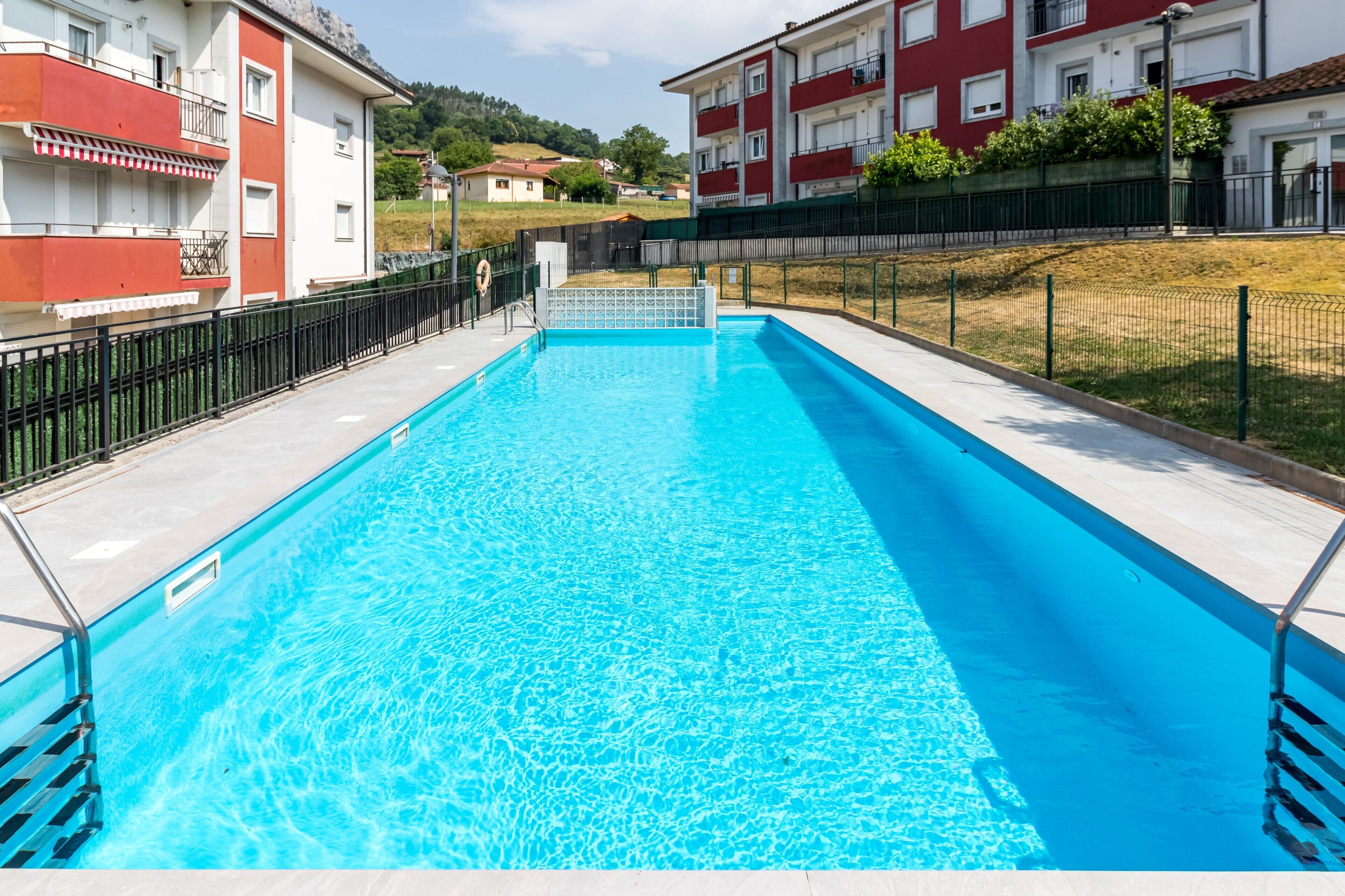 A rectangular swimming pool with clear blue water, surrounded by a black metal fence, and adjacent to a building with balconies.