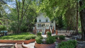 Una casa a due piani con un portico, circondata da un giardino in mattoni e alberi.