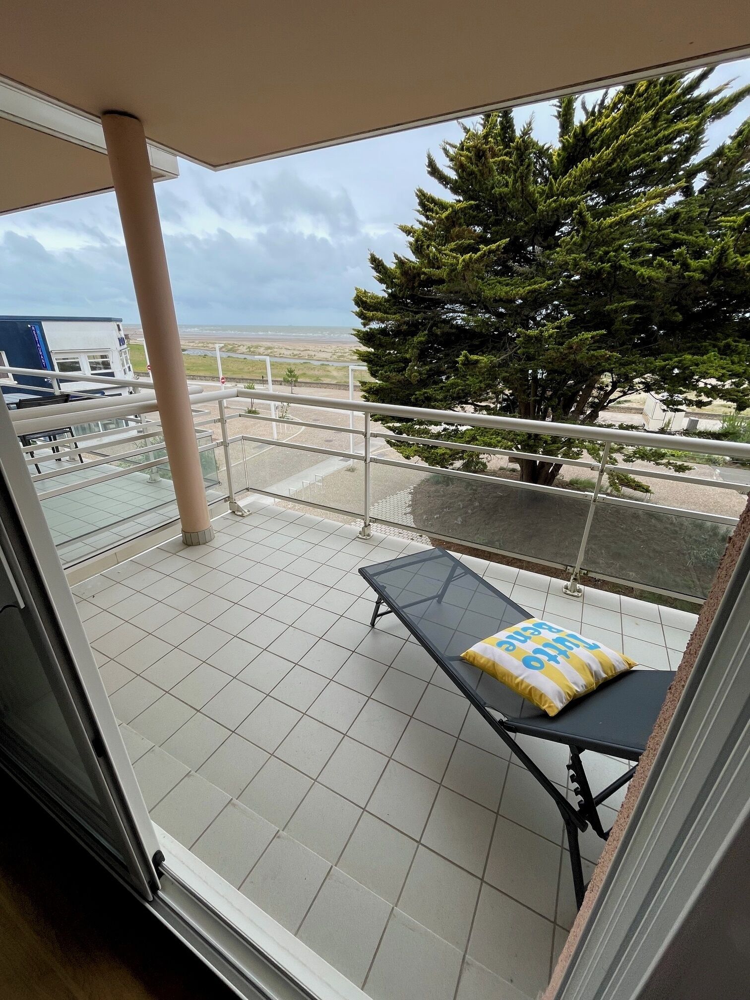 A balcony with a glass table and a cushion, a tiled floor, and a view of a beach and trees.