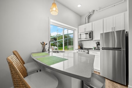 A modern kitchen with white cabinets, stainless steel appliances, a large island with seating, and a view of greenery outside.. 1 BR King Suite - Freshwater Inn - Bridgman, MI - Unit 1
