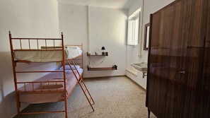 Ein Stockbett, ein kleines Regal mit einer Lampe, ein Waschbecken und ein Kleiderschrank in einem Raum mit Fliesenboden.