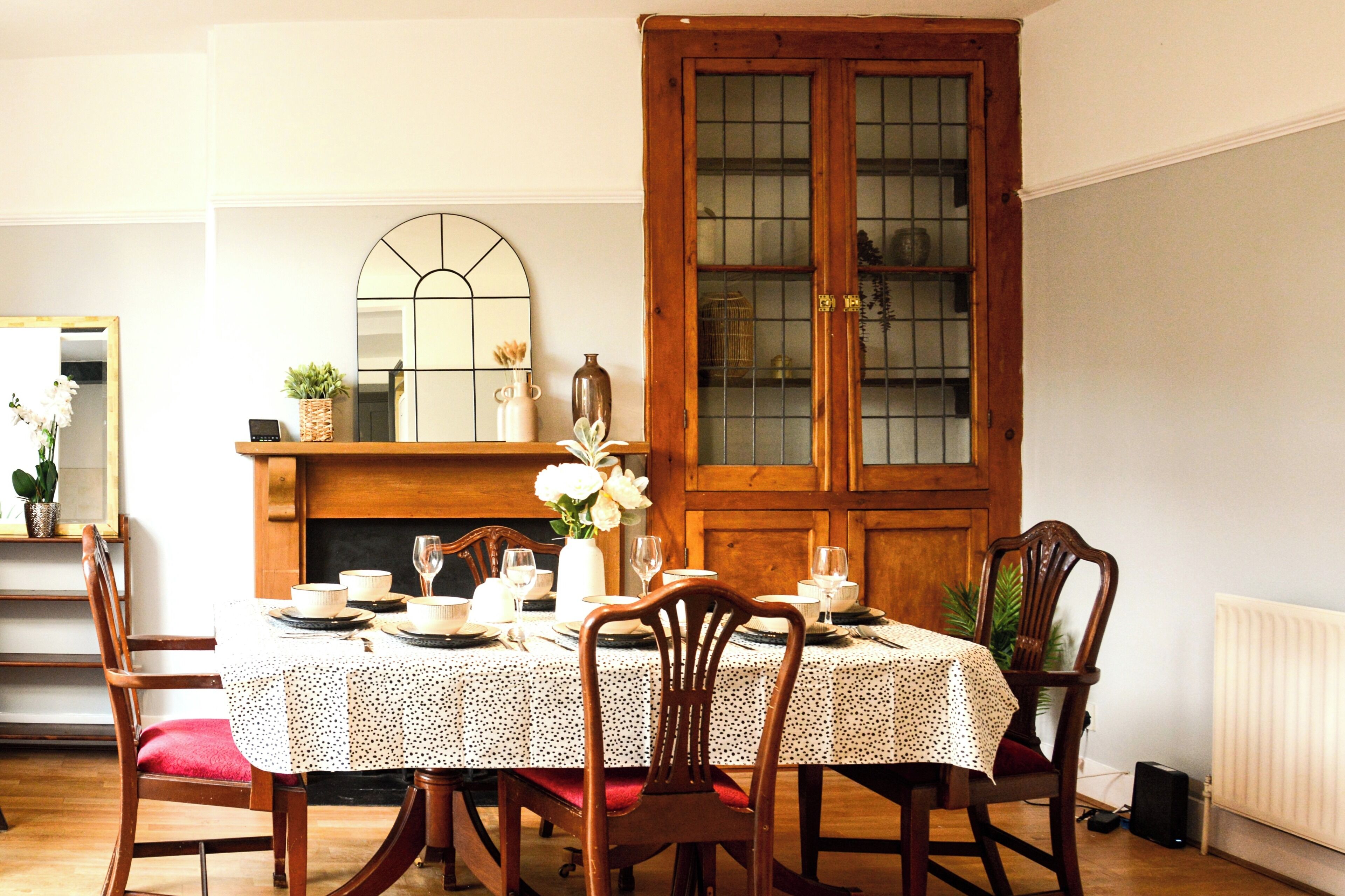 Un comedor con una mesa redonda dispuesta para cuatro personas, rodeada de sillas de madera. Se ve una chimenea con un espejo encima al fondo.