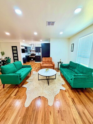 A living room with two green sofas, a round coffee table, and a kitchen in the background.