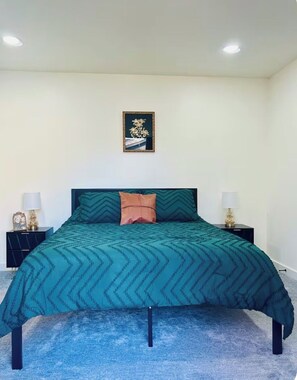 A bedroom with a bed, two bedside tables, a nightstand, a framed picture on the wall, and a carpeted floor.