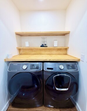 A laundry room with two Samsung washing machines, a wooden shelf above, and a water heater on the wall.