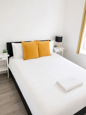 A neatly made bed with white and mustard pillows, a white nightstand with a lamp, and a small plant. - Hatfield | Entire House | Save Big for Long Stays (Hatfield)