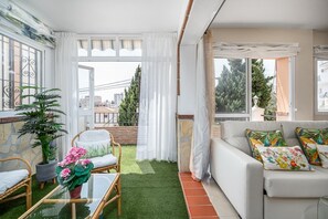 Un balcón con césped artificial, una silla de mimbre, un sofá blanco con cojines de colores y una planta en maceta.