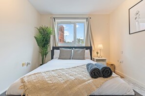 Un lit bien fait, recouvert d’une couverture beige et agrémenté de serviettes pliées, une lampe de chevet, une plante en pot et une fenêtre avec des rideaux.