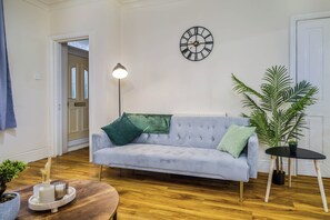 Ein Wohnzimmer mit Sofa, einer Lampe, einer Uhr, einer Pflanze und einem kleinen Tisch.