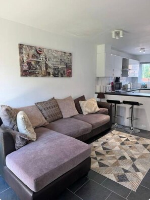 A modern living room with a sectional sofa, a kitchen area with white cabinets, and a geometric patterned rug. - Professionals | Entire House | 2BDR | Weekly Deals (London)
