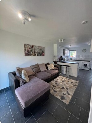 A modern living room with a sectional sofa, a kitchen area with a bar, and a patterned rug. - Professionals | Entire House | 2BDR | Weekly Deals (London)