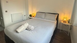 En snyggt bäddad säng med ett vitt täcke och kuddar, ett sängbord med en lampa och ett element på väggen.