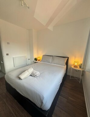 A neatly made bed with a white comforter and pillows, a bedside table with a lamp, and a radiator on the wall. - Professionals | Entire House | 2BDR | Weekly Deals (London)